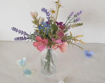 May include: A bouquet of artificial flowers in a clear glass vase. The bouquet includes lavender, daisies, pink and blue flowers. The flowers are arranged in a vase on a white surface.