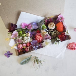 May include: A box filled with a variety of artificial flowers in shades of pink, purple, red, and white. The flowers are arranged in a loose, natural style, and the box is open, revealing the flowers inside.