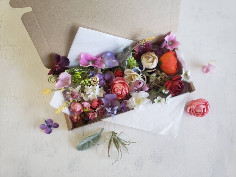 May include: A box filled with a variety of artificial flowers in shades of pink, purple, red, and white. The flowers are arranged in a loose, natural style, and the box is open, revealing the flowers inside.