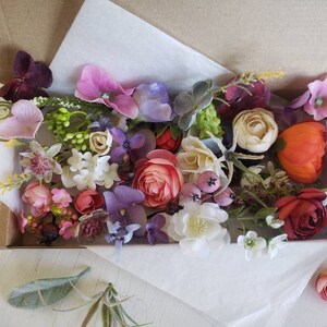 May include: A box filled with a variety of artificial flowers in shades of pink, purple, red, orange, and white. The flowers are arranged in a random pattern and include roses, carnations, and other types.