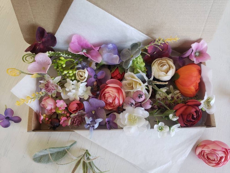 May include: A box filled with a variety of artificial flowers in shades of pink, purple, red, orange, and white. The flowers are arranged in a random pattern and include roses, carnations, and other types.