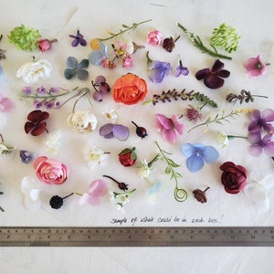 May include: A variety of artificial flowers and greenery in different colors, including pink, purple, blue, white, and green. The flowers are arranged on a white surface with a ruler in the bottom of the image. The text "Sample of what could be in each box." is written on the surface.