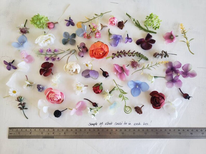 May include: A variety of artificial flowers and greenery in different colors, including pink, purple, blue, white, and green. The flowers are arranged on a white surface with a ruler in the bottom of the image. The text "Sample of what could be in each box." is written on the surface.