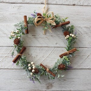 May include: A circular wreath adorned with cinnamon sticks, pine cones, and white berries, complemented by green foliage and lavender. The wreath is tied with twine and a raffia bow, offering a rustic, natural look. It hangs against a white wooden backdrop.