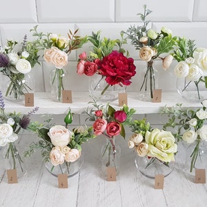 May include: Ten small glass vases filled with artificial flower arrangements. The vases are arranged in two rows on a white shelf. The flowers are a variety of colors, including white, pink, red, and green.