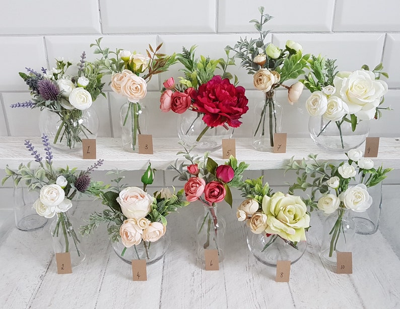 May include: Ten small glass vases filled with artificial flower arrangements. The vases are arranged in two rows on a white shelf. The flowers are a variety of colors, including white, pink, red, and green.