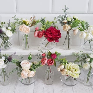 May include: Ten small glass vases filled with artificial flower arrangements. The vases are clear glass and round. The flowers are a variety of colors, including white, pink, red, and green.
