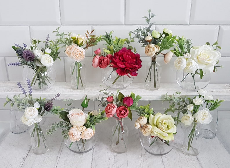 May include: Ten small glass vases filled with artificial flower arrangements. The vases are clear glass and round. The flowers are a variety of colors, including white, pink, red, and green.