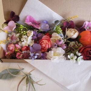 May include: A box filled with a variety of artificial flowers in shades of pink, purple, red, orange, and white. The flowers are arranged in a random pattern and include roses, carnations, and other types of flowers.