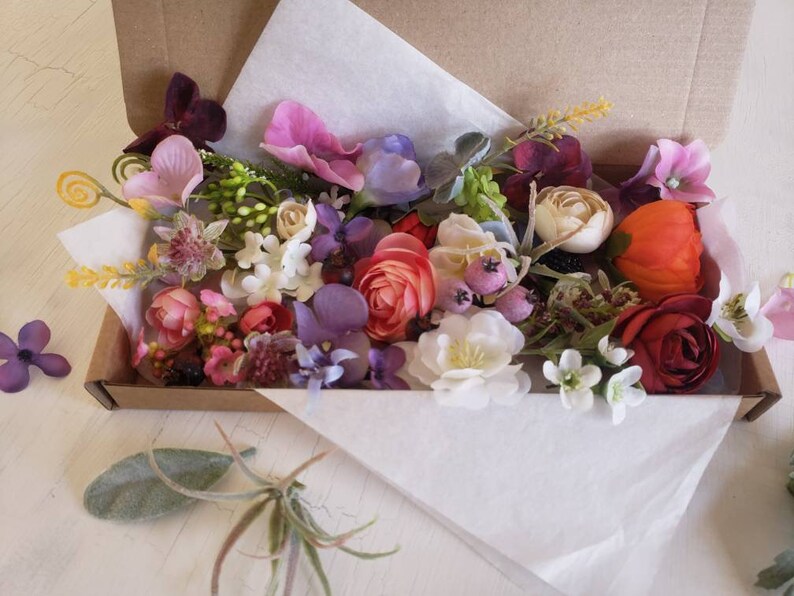 May include: A box filled with a variety of artificial flowers in shades of pink, purple, red, orange, and white. The flowers are arranged in a random pattern and include roses, carnations, and other types of flowers.