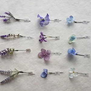 May include: Twelve different flower pins, including lavender and hydrangea, are displayed on a white surface. Each pin has a small paper tag with a number on it.