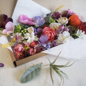 May include: A box filled with a variety of artificial flowers in shades of pink, purple, white, red, and yellow. The flowers are arranged in a loose, natural style and are perfect for crafting or decorating.
