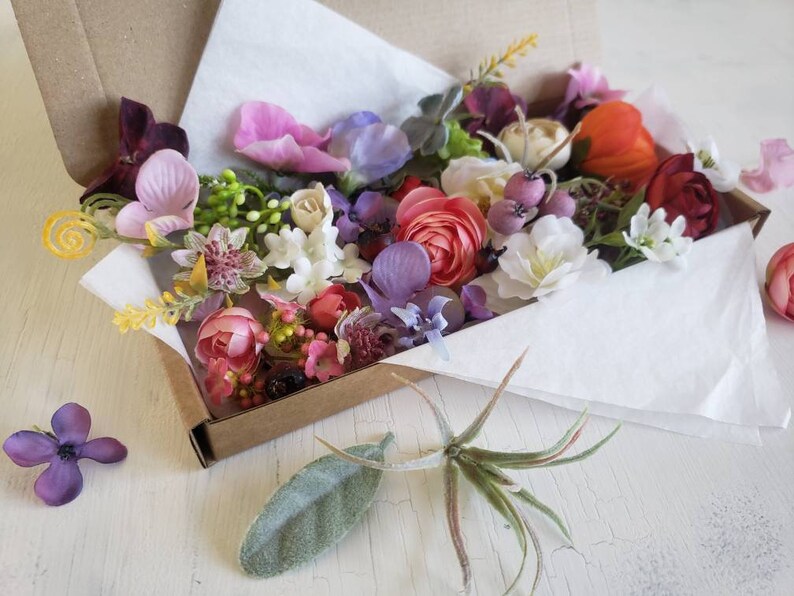 May include: A box filled with a variety of artificial flowers in shades of pink, purple, white, red, and yellow. The flowers are arranged in a loose, natural style and are perfect for crafting or decorating.