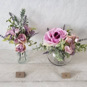 May include: Two floral arrangements in glass vases. The arrangement on the left is in a tall, clear vase and features pink and purple flowers with green foliage. The arrangement on the right is in a round, clear vase and features a large pink rose with smaller pink flowers and green foliage. Both arrangements have lavender sprigs.