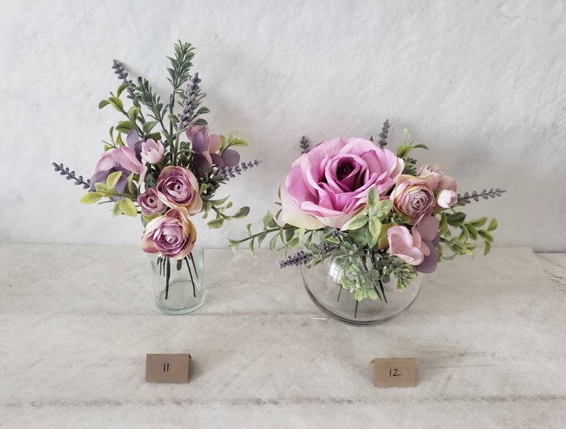 May include: Two floral arrangements in glass vases. The arrangement on the left is in a tall, clear vase and features pink and purple flowers with green foliage. The arrangement on the right is in a round, clear vase and features a large pink rose with smaller pink flowers and green foliage. Both arrangements have lavender sprigs.