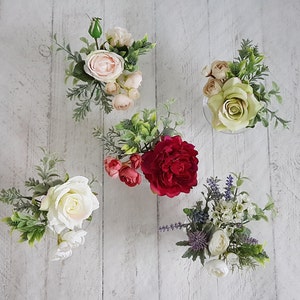 May include: Five small glass vases filled with artificial flowers. The vases are arranged in a row on a white wooden surface. The flowers are a variety of colors, including pink, red, white, and green.