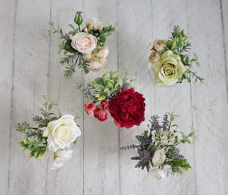 May include: Five small glass vases filled with artificial flowers. The vases are arranged in a row on a white wooden surface. The flowers are a variety of colors, including pink, red, white, and green.