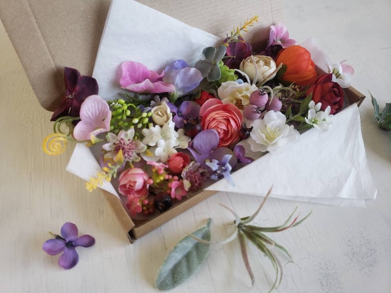 May include: A box filled with a variety of artificial flowers in shades of pink, purple, red, white, and yellow. The flowers are arranged in a loose, natural style and are perfect for crafting or decorating.