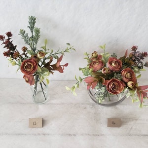 May include: Two floral arrangements in clear glass vases. The arrangement on the left has a mix of green foliage and red flowers. The arrangement on the right has a mix of green foliage and red flowers in a round vase. The arrangements are labeled with the numbers 13 and 14.