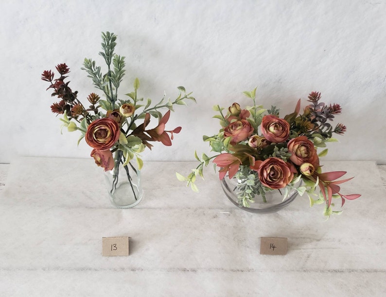 May include: Two floral arrangements in clear glass vases. The arrangement on the left has a mix of green foliage and red flowers. The arrangement on the right has a mix of green foliage and red flowers in a round vase. The arrangements are labeled with the numbers 13 and 14.