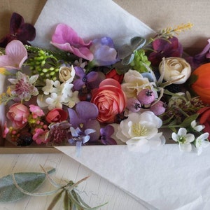 May include: A box filled with a variety of artificial flowers in shades of pink, purple, red, white, and green. The flowers are arranged in a random pattern and include roses, carnations, and other types.