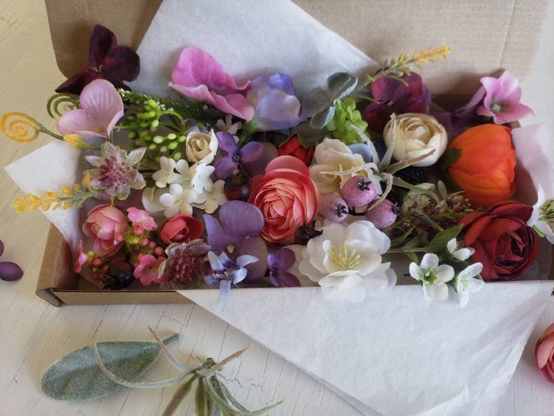 May include: A box filled with a variety of artificial flowers in shades of pink, purple, red, white, and green. The flowers are arranged in a random pattern and include roses, carnations, and other types.