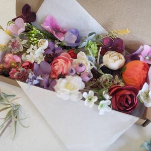 May include: A box filled with a variety of artificial flowers in shades of pink, purple, red, orange, and white. The flowers are arranged in a loose, natural style, and there are several different types of flowers, including roses, carnations, and daisies.
