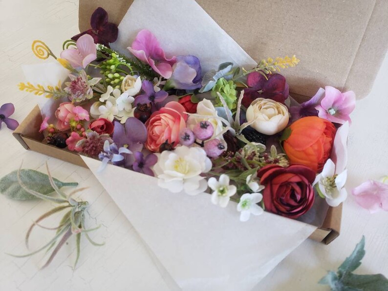 May include: A box filled with a variety of artificial flowers in shades of pink, purple, red, orange, and white. The flowers are arranged in a loose, natural style, and there are several different types of flowers, including roses, carnations, and daisies.