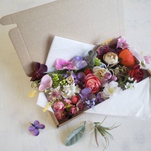 May include: A box filled with a variety of artificial flowers in shades of pink, purple, red, and orange. The flowers are arranged in a bouquet and are surrounded by green leaves.