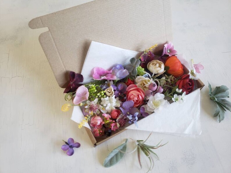 May include: A box filled with a variety of artificial flowers in shades of pink, purple, red, and orange. The flowers are arranged in a bouquet and are surrounded by green leaves.