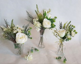 May include: Three white flower arrangements in clear glass vases. Each arrangement features white roses, white flowers, and green foliage.