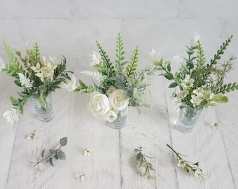 May include: Three small glass vases filled with white and green floral arrangements. The vases are arranged in a row on a white wooden surface.