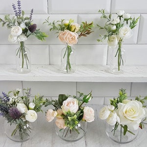 May include: Six glass vases filled with artificial flower arrangements. The vases are clear glass and the flowers are white, pink, and purple. The flowers are arranged in a variety of styles, some with a single type of flower and others with a mix of flowers.