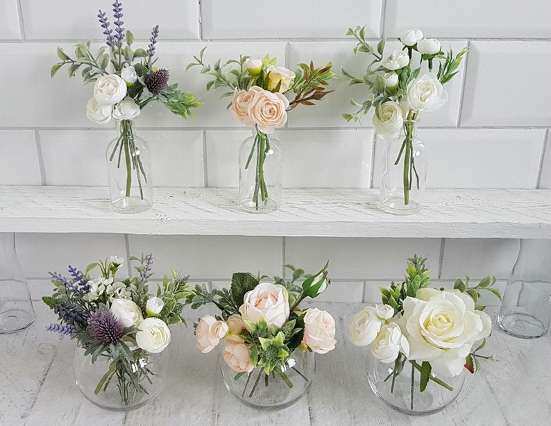 May include: Six glass vases filled with artificial flower arrangements. The vases are clear glass and the flowers are white, pink, and purple. The flowers are arranged in a variety of styles, some with a single type of flower and others with a mix of flowers.