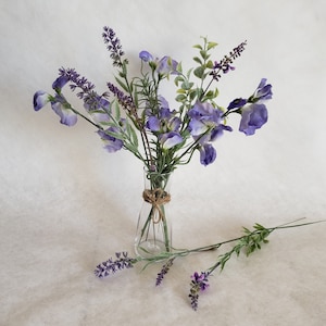 May include: A bouquet of purple and green flowers in a clear glass vase with a brown twine bow. The flowers are arranged in a vase on a white background.