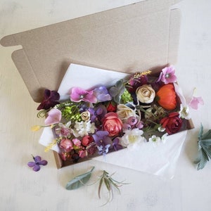 May include: A box filled with a variety of artificial flowers in shades of pink, red, purple, and white. The flowers are arranged in a loose, natural style and include roses, carnations, and other blooms.