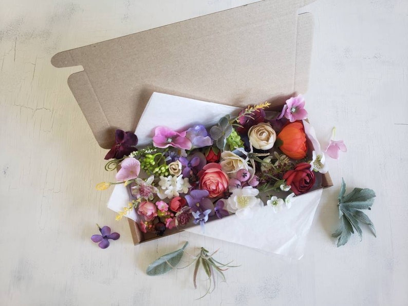 May include: A box filled with a variety of artificial flowers in shades of pink, red, purple, and white. The flowers are arranged in a loose, natural style and include roses, carnations, and other blooms.