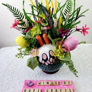 May include: A spring-themed floral arrangement in a blue ceramic pot with a white bunny rabbit with pink ears and paws. The arrangement includes yellow, pink, and purple flowers, green ferns, and yellow and pink Easter eggs. The pot is sitting on a pink surface with the words "Wings of Hope Crafts" in wooden letter tiles.