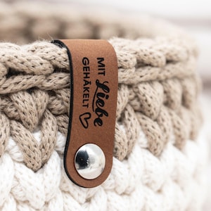 Könnte beinhalten: Nahaufnahme eines handgefertigten Häkelkorbs in Creme, Beige und Weiß. Ein braunes Lederetikett ist mit den Worten "Mit Liebe Gehäkelt" und einem Herzsymbol befestigt. Der Korb ist aus dickem Garn gefertigt.