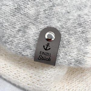 May include: A dark grey rectangular leather tag attached to a light grey and white knit fabric. The tag features a silver metal rivet at the top, a black anchor icon, and the black printed text "EINZEL ST&Uuml;CK" centred on the surface.