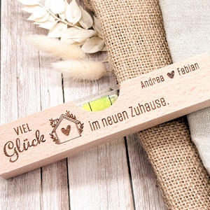 Könnte beinhalten: Ein Holz-Wasserwaage mit Gravur und einem Haus-Motiv. Der Text lautet "Viel Glück im neuen Zuhause" und "Andrea ♥ Fabian". Die Wasserwaage hat eine Libelle mit grüner Flüssigkeit.