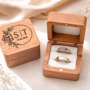 Könnte beinhalten: Eine quadratische Ringbox aus Holz mit Klappdeckel, der mit einem lasergravierten Blumenkranz, den Buchstaben S und T sowie dem Datum 13.06.2026 versehen ist. Die geöffnete Box zeigt ein weißes Samtfutter mit zwei silberfarbenen Metallringen.