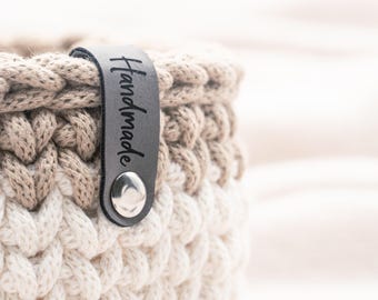 May include: A round crochet basket featuring a two-tone design with a beige upper section and a cream lower section. A dark grey leather tag is attached to the rim with a silver metal rivet, featuring the word Handmade embossed in black text.