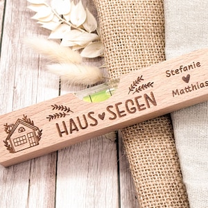 Könnte beinhalten: Ein Holz-Wasserwaage mit den Worten "HAUS SEGEN" und einer eingravierten Hausillustration. Die Wasserwaage trägt auch die Namen "Stefanie" und "Matthias". Sie hat eine Blasenampulle mit grüner Flüssigkeit.