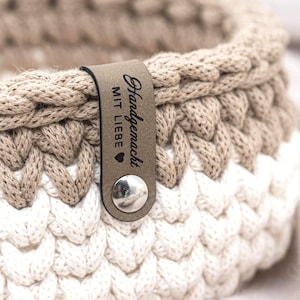 Könnte beinhalten: Nahaufnahme eines handgefertigten Häkelkorbs in Beige und Weiß. Der Korb hat ein geflochtenes Design und ein Lederetikett mit den Worten "Handgemacht MIT LIEBE" und einem Herzsymbol. Das Etikett ist mit einer silbernen Niete befestigt.