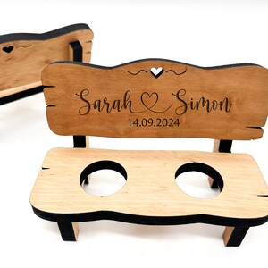 May include: A wooden bench with two cup holders, personalised with the names "Sarah & Simon" and the date "14.09.2024". The bench is painted black and has a heart cutout on the back.