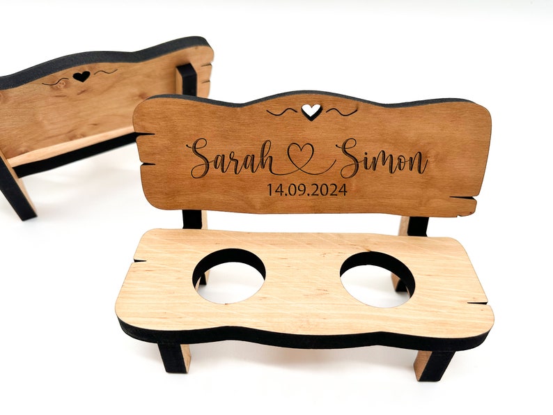 May include: A wooden bench with two cup holders, personalised with the names "Sarah & Simon" and the date "14.09.2024". The bench is painted black and has a heart cutout on the back.