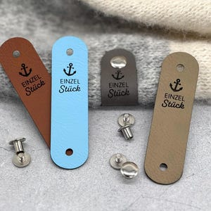 May include: Four rectangular faux leather clothing labels in brown, light blue, dark grey, and tan. Each label features a black anchor icon and the text "EINZEL St&uuml;ck" printed in black. The labels include pre-punched holes and separate metal rivets.