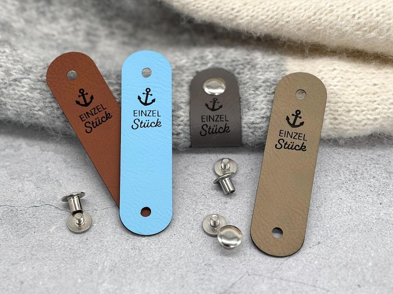 May include: Four rectangular faux leather clothing labels in brown, light blue, dark grey, and tan. Each label features a black anchor icon and the text "EINZEL St&uuml;ck" printed in black. The labels include pre-punched holes and separate metal rivets.