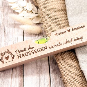 Könnte beinhalten: Ein Holz-Wasserwaage mit Libelle, graviert mit Text und einer Hausillustration. Der Text enthält Namen und einen deutschen Satz. Die Wasserwaage ist hellbraun und liegt auf einer Holzoberfläche.